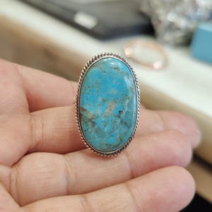 May include: A silver ring with a large oval turquoise stone set in a silver bezel. The stone has a blue and green color with brown and white veins.