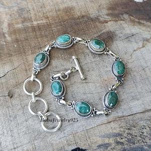 May include: A silver bracelet with a toggle clasp features oval-shaped green gemstones set in silver. The bracelet has a delicate, intricate design.