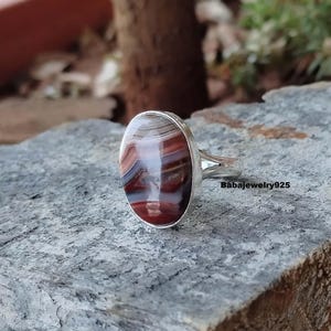 May include: Oval ring with a polished agate stone in red, brown, and white bands. The stone is set in a silver-coloured metal band. The ring is displayed on a grey stone surface. The text "Babajewelry925" is visible.