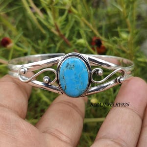 May include: A silver cuff bracelet featuring a large, oval turquoise stone at the center. The bracelet has decorative swirls and the text "BABA JEWELRY925". The turquoise stone is a vibrant blue color.