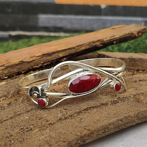 May include: A silver bangle bracelet with a red gemstone and two smaller red gemstones. The bracelet has a delicate, intricate design.