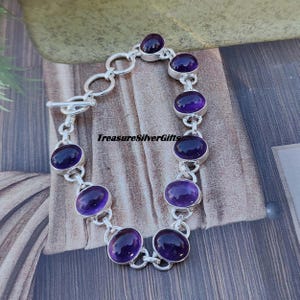 K&ouml;nnte beinhalten: Ein silbernes Kettenarmband mit acht ovalen Amethyst-Edelsteinen. Das Armband hat einen Schiebeverschluss.