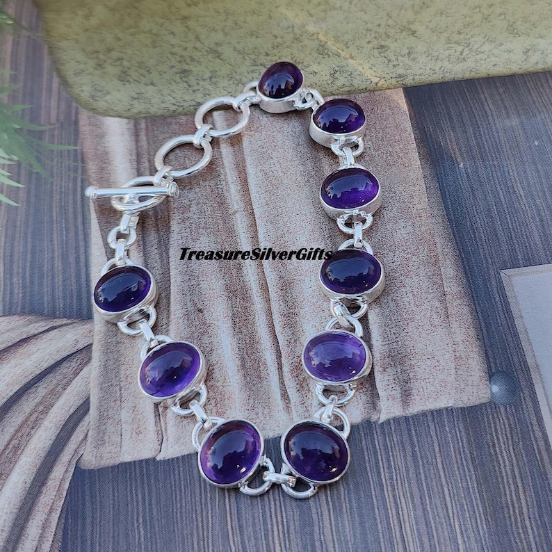 K&ouml;nnte beinhalten: Ein silbernes Kettenarmband mit acht ovalen Amethyst-Edelsteinen. Das Armband hat einen Schiebeverschluss.