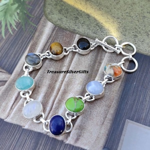 May include: A silver bracelet with a toggle clasp features nine different gemstone cabochons. The stones are set in silver and include tiger's eye, black onyx, labradorite, aquamarine, blue lace agate, moonstone, turquoise, and lapis lazuli.