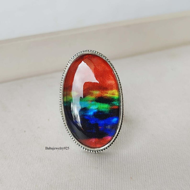 May include: A silver ring with an oval-shaped, rainbow-colored glass stone. The stone has a red, orange, yellow, green, blue, and purple color scheme.