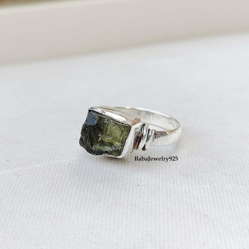 May include: A silver ring with a dark green gemstone. The ring has a simple band with a square setting for the stone.