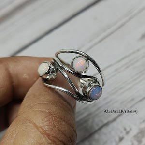 May include: A silver ring with three opal gemstones. The ring has a unique design with a curved band and three round opal stones in white and blue hues. The text "925JEWELRYBABAJ" is visible.