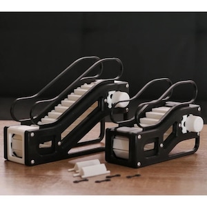 May include: Two miniature black and white escalators are displayed on a wooden surface. The escalators have a detailed design with steps, handrails, and mechanical components. Additional white and black pieces are scattered nearby.