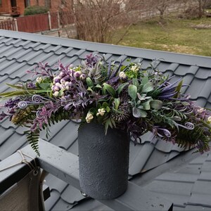 Op de afbeelding: Een boeket van paarse en witte kunstbloemen in een grijze, getextureerde vaas. De bloemen zijn in een watervalvorm gerangschikt en zijn gemaakt om op verschillende soorten bloemen te lijken.