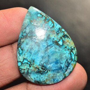 May include: A teardrop-shaped gemstone, predominantly turquoise, with detailed brown and black veining. The polished surface catches the light, highlighting the stone's natural beauty. The gemstone is held in a hand.