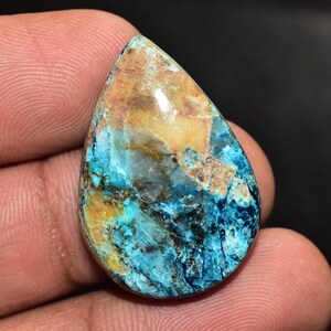 May include: A teardrop-shaped gemstone with a polished surface. The stone features a blend of colours, including shades of blue, brown, and gold. The gemstone is held in a hand against a dark background.