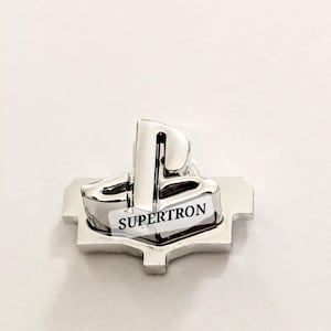 May include: A silver PlayStation logo button with the word "SUPERTRON" printed on it.