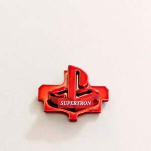 May include: A red, metallic pin with the text "SUPERTRON". The pin has a distinctive shape with a central design and a surrounding frame. The pin is set against a white background.