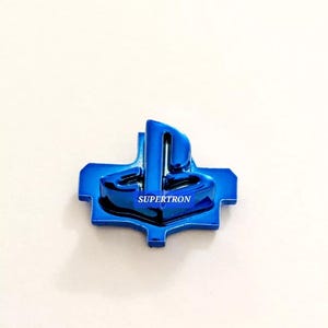 May include: A blue PlayStation button with a metallic finish. The button has a raised PlayStation logo and the word "SUPERTRON" printed on it.
