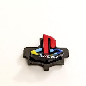May include: A black emblem with a red, blue, and yellow PlayStation logo and the word "SUPERTRON" in white. The emblem is set against a white background.
