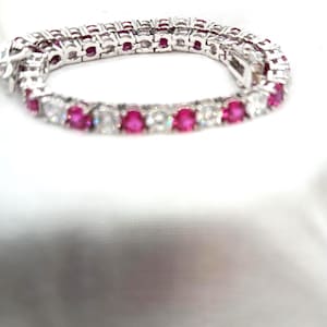 May include: A silver bracelet with alternating pink and clear gemstones. The bracelet is displayed on a white surface, showcasing the sparkle of the gems. The gemstones are round and set closely together, creating a continuous line of color and brilliance.