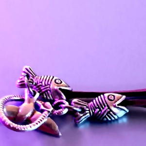 May include: Silver-toned metal charm bracelet with fish skeleton and snake designs. The bracelet features two fish skeleton charms and a snake charm, all connected by a dark brown cord. The background is a solid purple color.