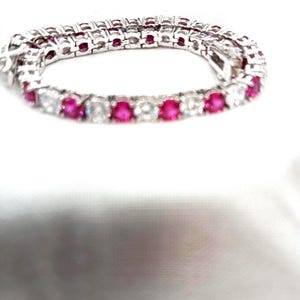 May include: A tennis bracelet with alternating round-cut, bright pink and clear gemstones set in a silver-colored metal. The bracelet is displayed on a white surface.