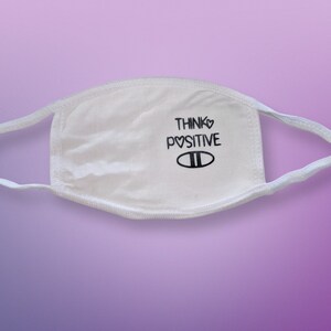 May include: White face mask with black text that reads "Think♥Positive" and a black heart symbol. The mask has two black lines below the text.