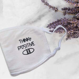 May include: White face mask with the text "Think ♥ Positive" and a heart symbol in the text. The mask has a white elastic band.