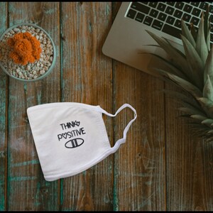 May include: White face mask with the text "Think Positive" and a heart symbol. The mask is on a wooden surface with a potted cactus and a pineapple.