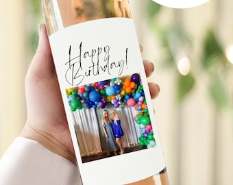 Puede incluir: Una etiqueta de botella de vino blanco con el texto "Happy Birthday!" en letra cursiva negra. La etiqueta presenta una foto de dos personas de pie frente a un arco de globos de colores.