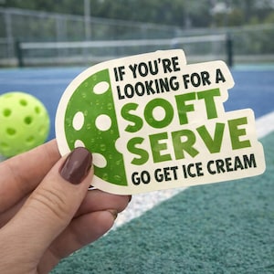 May include: A sticker with the text "IF YOU'RE LOOKING FOR A SOFT SERVE GO GET ICE CREAM" is held in front of a pickleball and a pickleball court. The sticker has a green and white design. The pickleball is yellow-green.