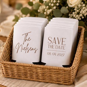 May include: A woven basket filled with white can coolers. One stack reads "The Nielsens" in brown script, and another says "SAVE THE DATE 08.04.2027". The basket sits on a beige tablecloth, with candles and flowers in the background.