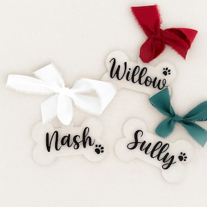 May include: Bone-shaped pet tags with names "Willow", "Nash", and "Sully" in black script, each with a paw print. White, red, and teal ribbons are tied in bows. The tags are on a white background.