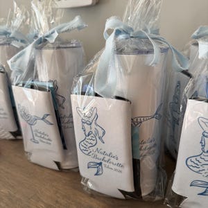May include: White insulated tumblers and can coolers, presented as gifts. Each tumbler is wrapped in clear cellophane and tied with a pale blue ribbon. The can coolers feature a blue mermaid design and the text "Natalie's Bachelorette Tulum 2026."
