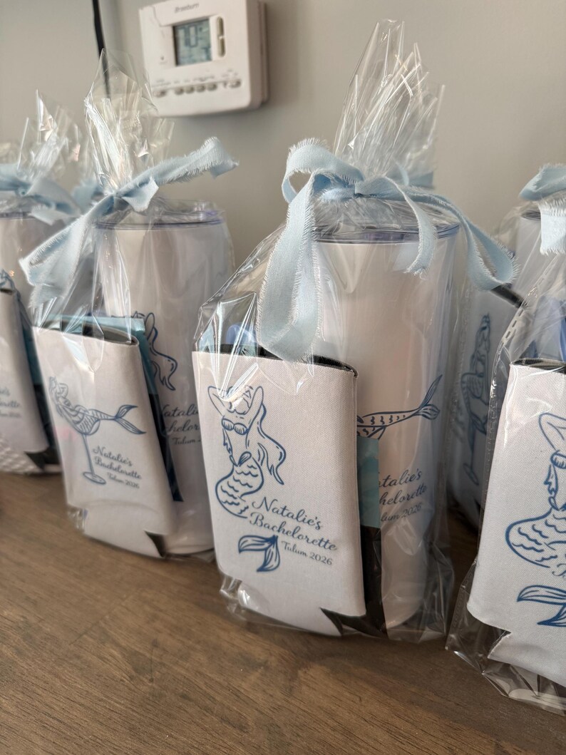 May include: White insulated tumblers and can coolers, presented as gifts. Each tumbler is wrapped in clear cellophane and tied with a pale blue ribbon. The can coolers feature a blue mermaid design and the text "Natalie's Bachelorette Tulum 2026."
