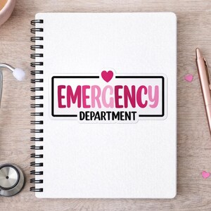May include: A white spiral-bound notebook with a sticker that reads "EMERGENCY DEPARTMENT" in pink and black. A pink stethoscope and pen are on the wooden surface. Pink heart-shaped confetti is scattered around the pen.