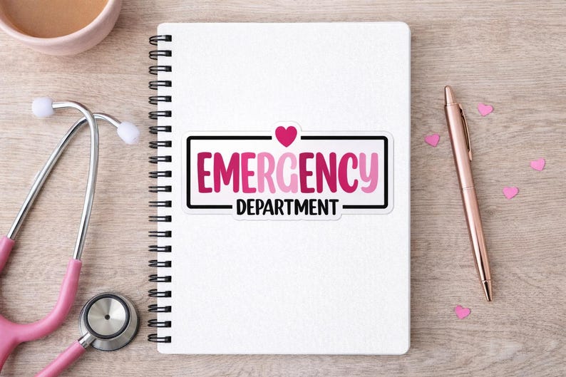 May include: A white spiral-bound notebook with a sticker that reads "EMERGENCY DEPARTMENT" in pink and black. A pink stethoscope and pen are on the wooden surface. Pink heart-shaped confetti is scattered around the pen.