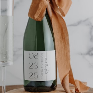 May include: A green champagne bottle with a white label featuring the date "08 23 25" and the text "save the date" with the names "Gabriella & Michael". The bottle is adorned with a brown ribbon.