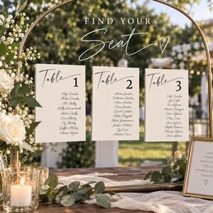 May include: A gold metal arch frame displays three white rectangular signs with black text reading "FIND YOUR SEAT" and "Table 1," "Table 2," and "Table 3" with guest names. A small framed sign reads "GOOD PEOPLE BEAUTIFUL PLACES UNFORGETTABLE NIGHTS."