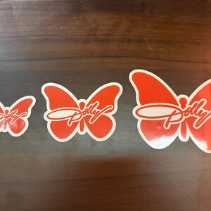 May include: Three orange butterfly-shaped stickers arranged in a row from smallest to largest. Each sticker features a white outline and the word Dolly written in a white cursive script across the center of the butterfly wings.