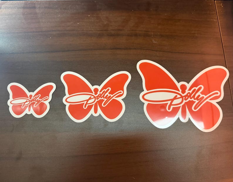 May include: Three orange butterfly-shaped stickers arranged in a row from smallest to largest. Each sticker features a white outline and the word Dolly written in a white cursive script across the center of the butterfly wings.