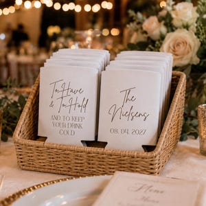 May include: A collection of white foam drink sleeves arranged in a woven basket. The sleeves feature black printed text that reads To Have & To Hold AND TO KEEP YOUR DRINK COLD on one side and The Nielsens 08. 04. 2027 on the other side.