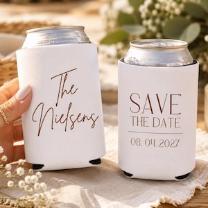 May include: Two white can coolers with brown text. One says "The Nielsens" and the other says "SAVE THE DATE" with the date "08.04.2027". The coolers are on a light-colored surface.