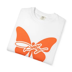 May include: A white short-sleeved t-shirt featuring a large orange butterfly graphic on the front. The word Dolly is written in a white cursive script across the center of the butterfly. The inner neck label displays the text Comfort Colors.