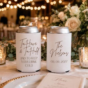 May include: Two white foam can coolers with black text. The left cooler reads To Have & To Hold AND TO KEEP YOUR DRINK COLD. The right cooler reads The Nielsens 08. 04. 2027. Both feature a silver metal top rim and a cylindrical shape for beverage cans.