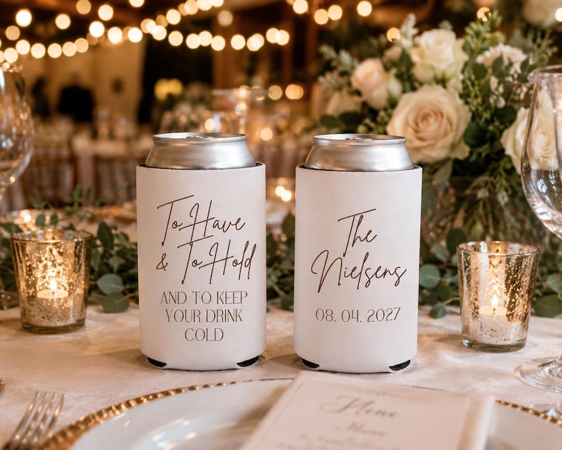 May include: Two white foam can coolers with black text. The left cooler reads To Have & To Hold AND TO KEEP YOUR DRINK COLD. The right cooler reads The Nielsens 08. 04. 2027. Both feature a silver metal top rim and a cylindrical shape for beverage cans.