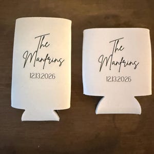 May include: Two white foam beverage insulators of different heights, each printed with the black text The Manfring and the date 12.13.2026 in a script and sans-serif font combination, designed to hold cans or bottles securely.