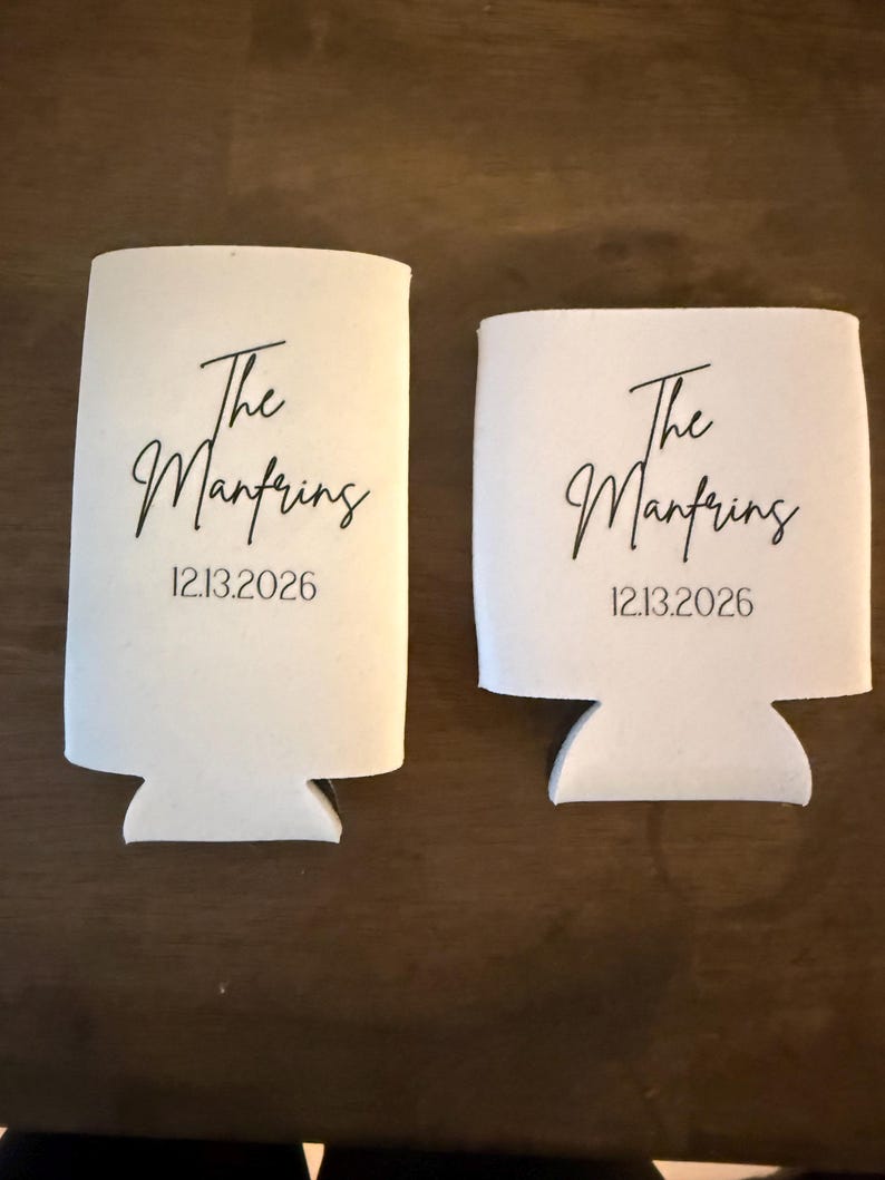 May include: Two white foam beverage insulators of different heights, each printed with the black text The Manfring and the date 12.13.2026 in a script and sans-serif font combination, designed to hold cans or bottles securely.