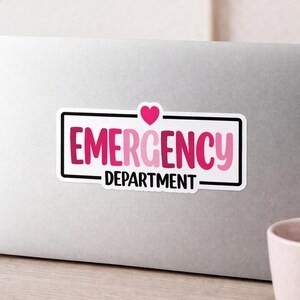 May include: A rectangular sticker with the words "EMERGENCY DEPARTMENT" in pink and white, with a pink heart above the word "EMERGENCY". The sticker has a black border and is on a silver laptop.