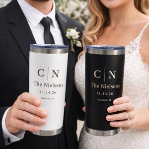 May include: Two stainless steel tumblers, one white and one black, each featuring a clear lid and custom engraving. The text reads C | N, The Nielsens, 11.14.26, and Chicago, IL. The tumblers have a cylindrical shape with a metallic rim.