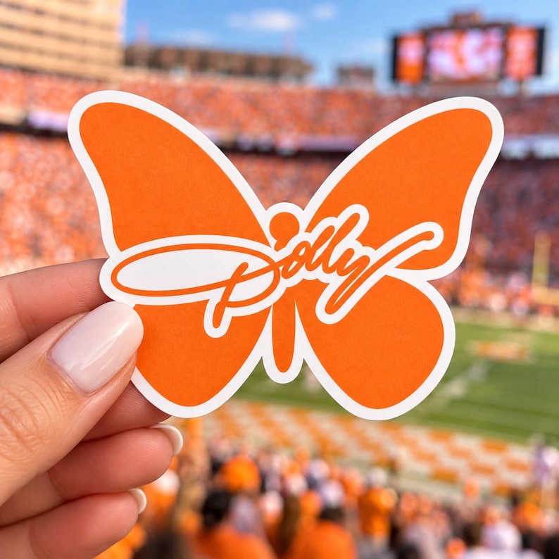 May include: A die-cut sticker shaped like a butterfly with bright orange wings and a white border. The word Dolly is written across the center of the butterfly in a white cursive script font. The sticker has a smooth, matte finish.