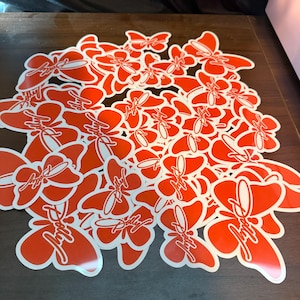 May include: A collection of numerous bright orange butterfly-shaped stickers scattered together. Each sticker features a white outline and the word Dolly written in a white cursive script across the center of the butterfly wings.