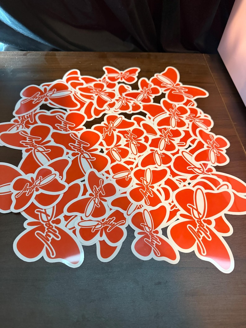 May include: A collection of numerous bright orange butterfly-shaped stickers scattered together. Each sticker features a white outline and the word Dolly written in a white cursive script across the center of the butterfly wings.