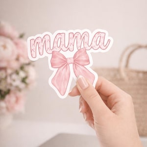 May include: A pink sticker with the word "mama" in a cursive font, adorned with a bow. The sticker is held in front of a blurred background with a laptop and flowers.